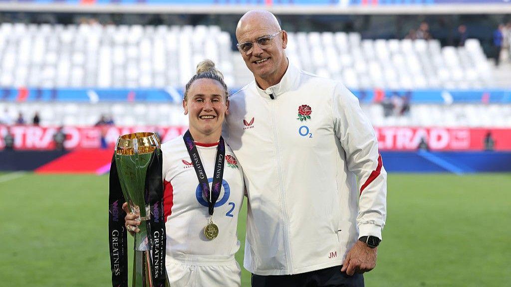Meg Jones (left), wearing a white England kit, holds the Six Nations trophy and poses with head coach John Mitchell, wearing a white England tracksuit top
