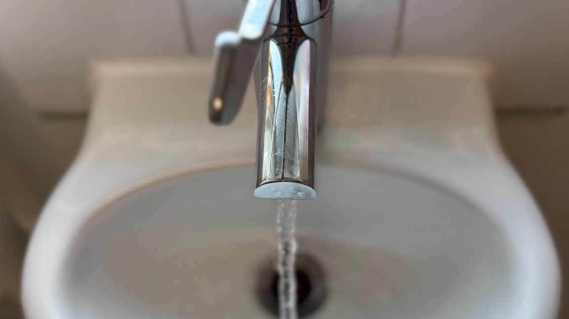 A shiny silver tap expels a strong flow of water into a white sink. The handle of the tap is over the middle of the outlet, and the water is pouring out directly over the sinkhole. The basin is attached to a white tiled wall.