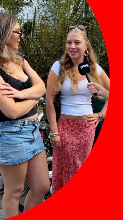 A woman with a white T-shirt and red skirt smiles in front of a mic. She's in the street with her friend