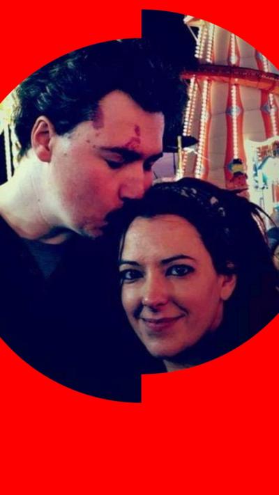 A woman with long dark hair smiles at the camera. On her right, a man with dark hair leans down and kisses her on the head. An illuminated helter-skelter is visible in the background.