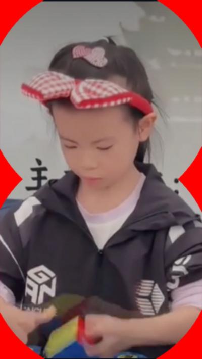 A six-year-old girl stood at desk competing at a Rubik's Cube competition. She is holding a Rubik's Cube.