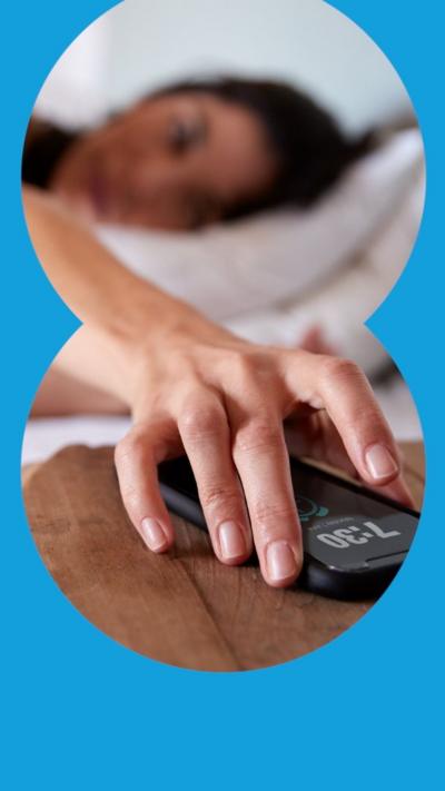 A woman reaches over to her phone on her bedside table from her bed. She has just woken up and it is light in the room.