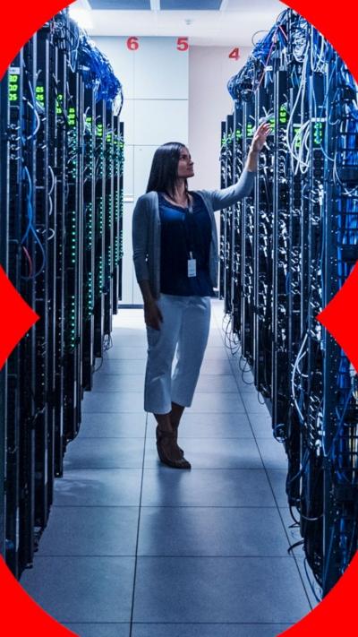 A woman with long dark hair stands in the middle of a corridor filled with towers of computer servers and wires that are taller than her