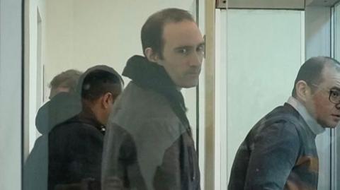A screengrab from a video shows a man in a hoodie standing with short hair and a jacket and looking at the camera