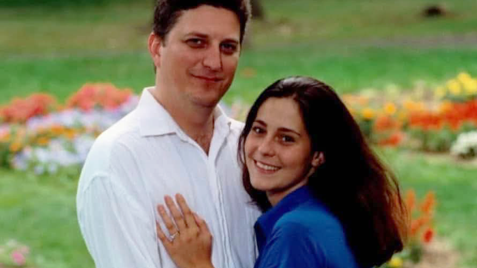 John Resta is on the left, he has dark hair and is wearing a white linen shirt and his wife Sylvia is on the right, with dark long hair and a blue short-sleeved shirt. She is pressed against him, her left hand on his chest. It has a wedding ring on it. They are both smiling. Behind them is grass with flowers.