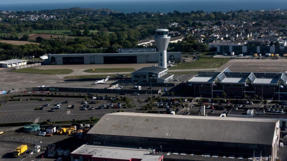 An aerial photo shows an airport control tower from a distance. It's behind a terminal building and a car park with lots of free spaces. Beside it is the tarmac taxiway and green grass spaces. In the background there are lots of trees, homes, and the sea on the horizon.