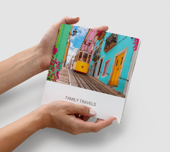 Hands holding a square custom photo book titled "FAMILY TRAVELS" with a picture of a yellow streetcar.